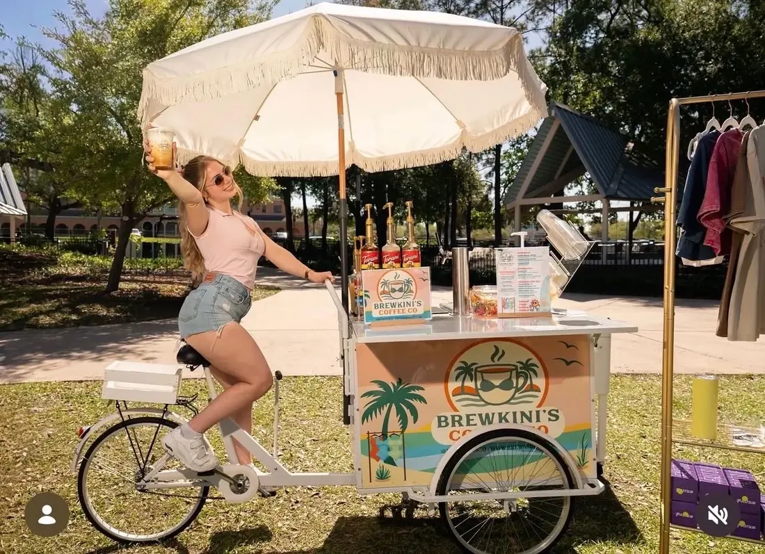 A BrewKini’s barista riding the Coffee Bar Trike under a fringed umbrella